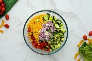 Bowl of lettuce topped with diced cucumbers, red onions, sliced tomatoes, and corn.