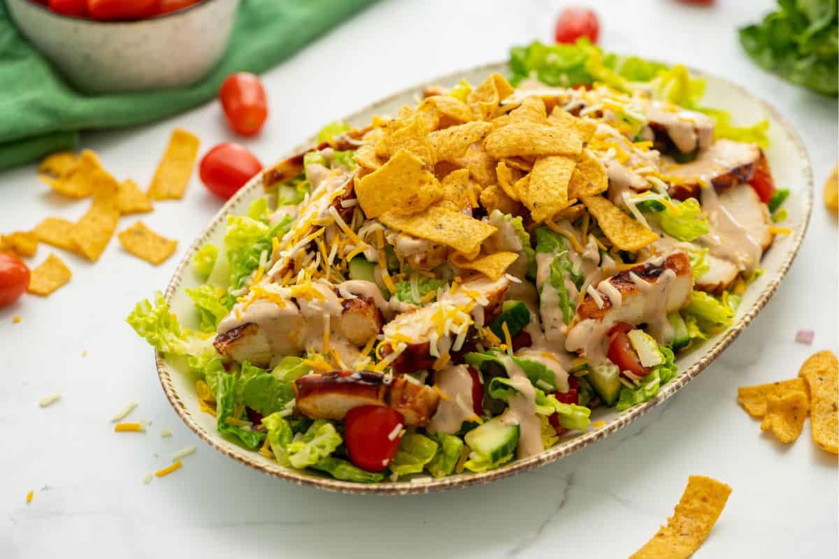 Bed of lettuce topped with BBQ Chicken, shredded cheese, corn chips, and BBQ Ranch Dressing with tomatoes in background.