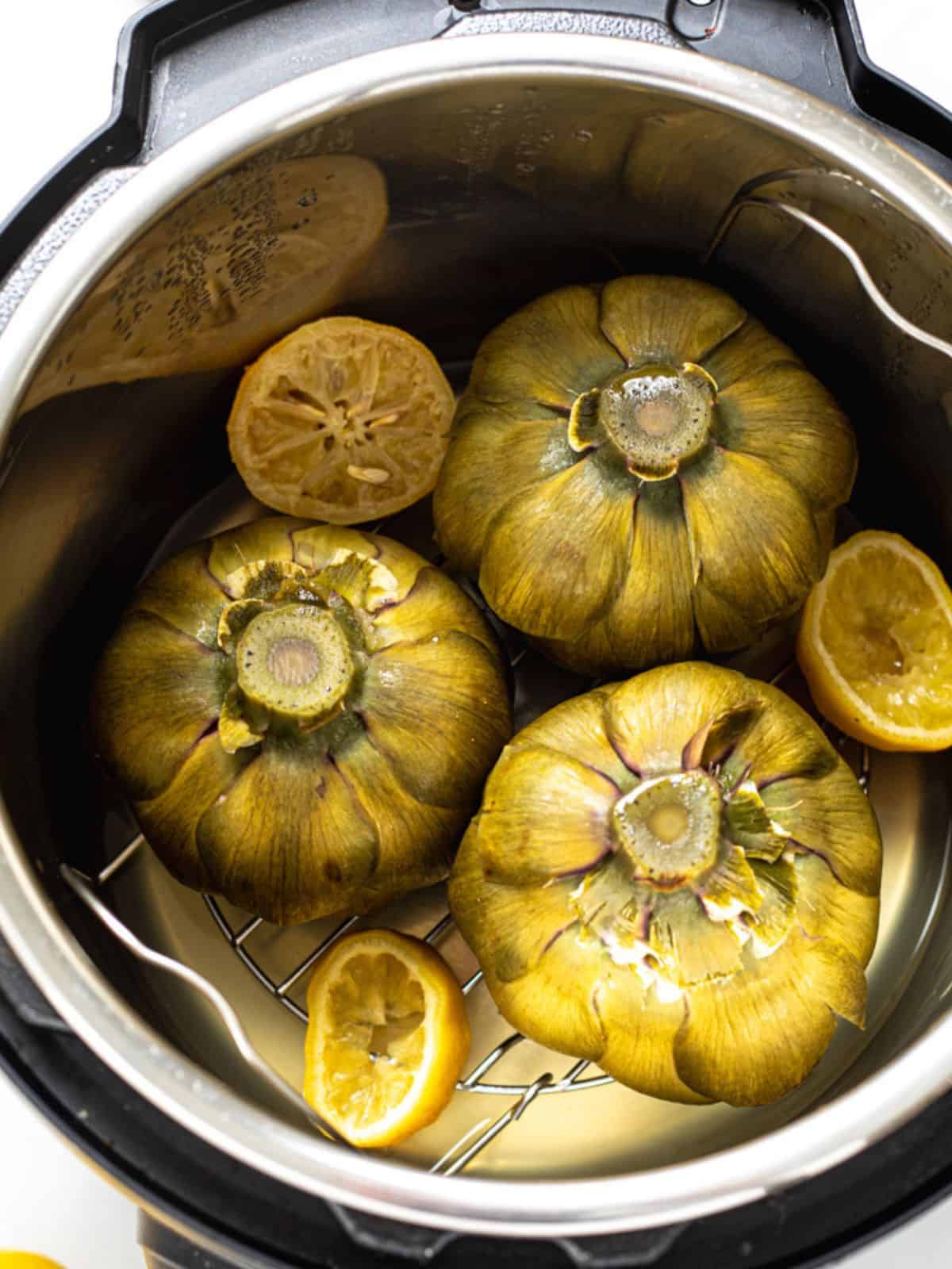 Three pressure cooked whole artichokes inside inner pot on metal trivet with sliced lemon.