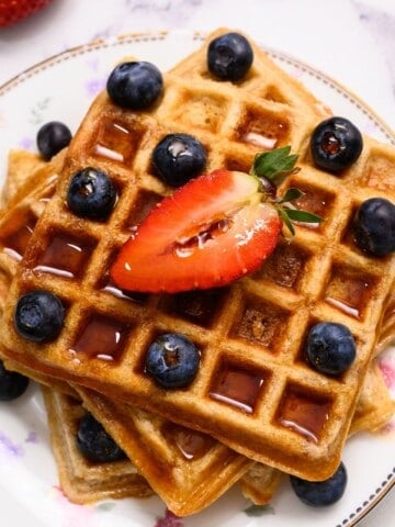 3 whole wheat waffles on white plate topped with berries and maple syrup.