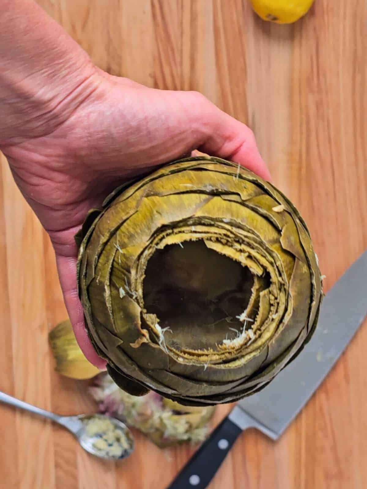 Whole artichoke that has been hollowed out with spoon and choke discarded on wooden cutting board in background.