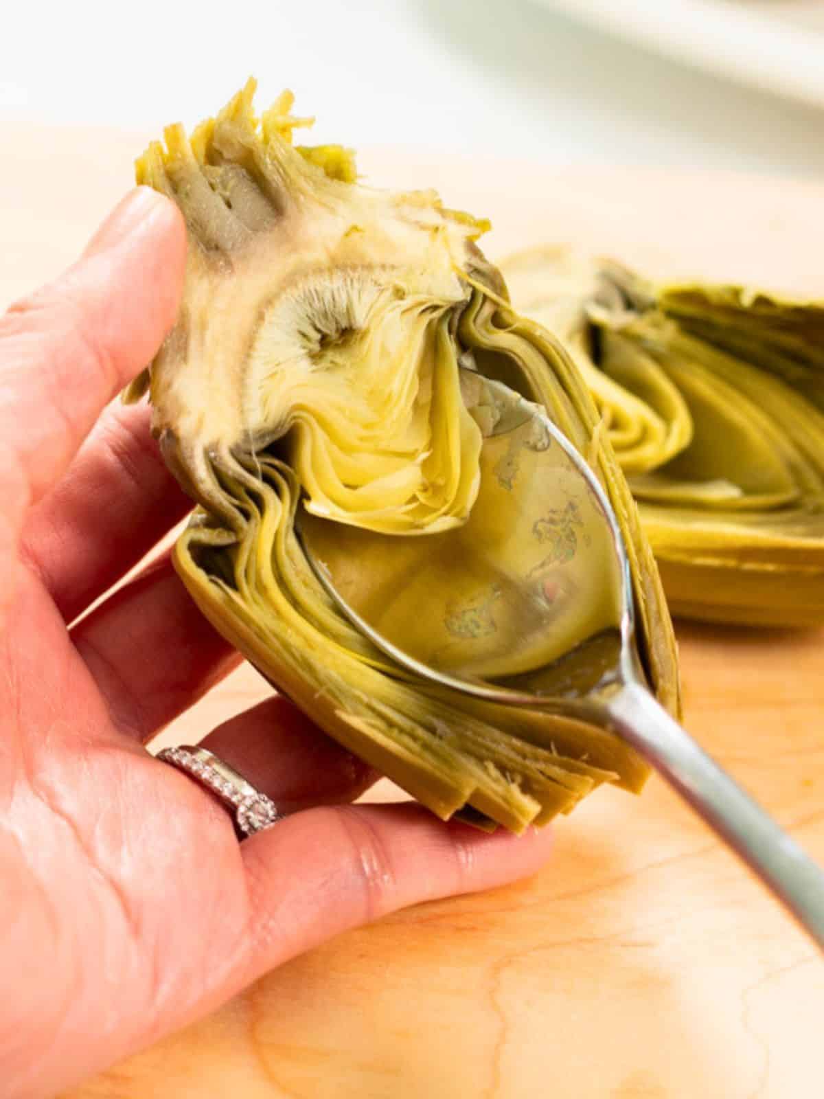 Artichoke cut in half and spoon removing thistle from heart of artichoke.