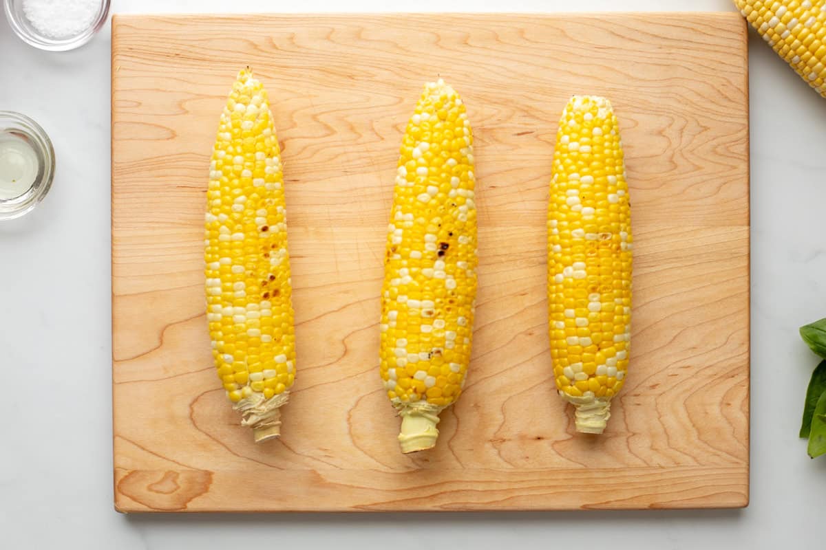 Three ears of grilled corn on wooden cutting board.