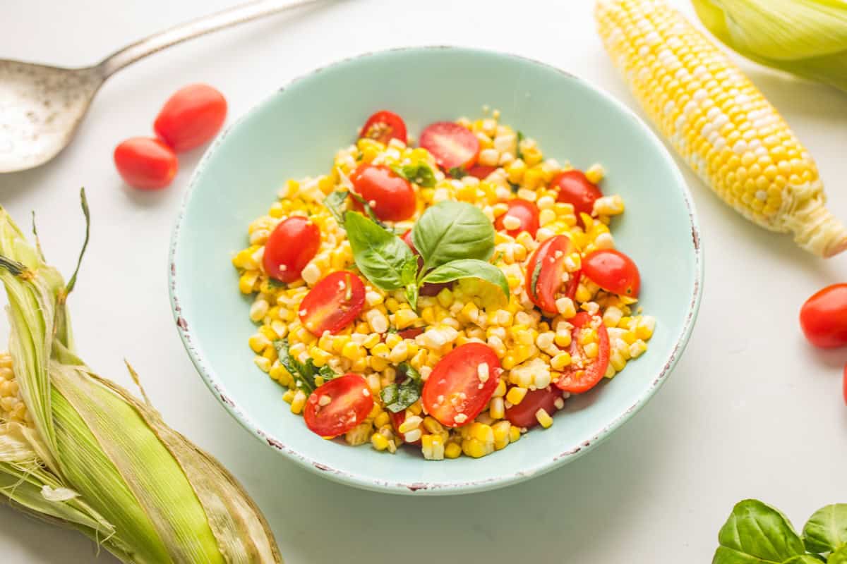 Fresh corn and tomato salad with basil in blue bowl with corn on the cob and grape tomatoes on the counter.