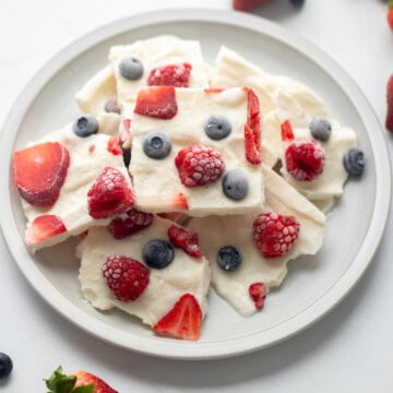 Broken frozen yogurt bark topped with berries on light gray plate surrounded by fresh berries.