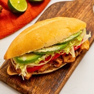Chicken torta on wooden cutting board, topped with sliced tomatoes, cabbage, and avocado slices.