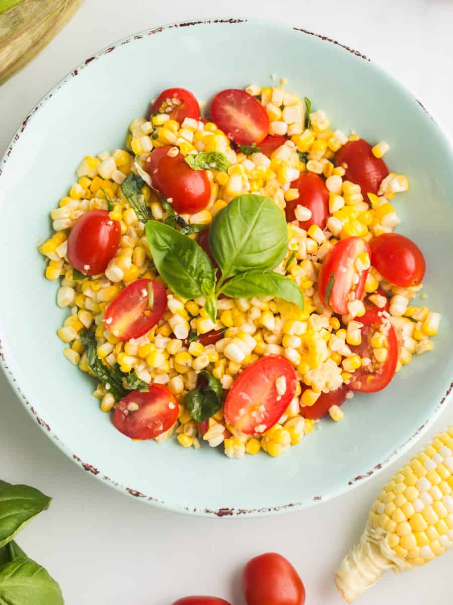 Fresh corn and tomato salad with basil in blue bowl with corn on the cob and grape tomatoes on the counter.