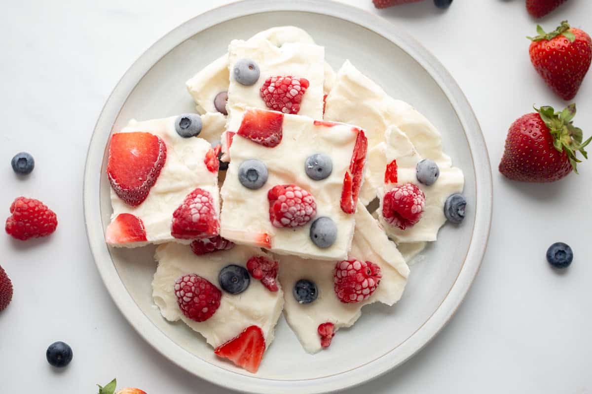 Broken frozen yogurt bark topped with strawberries, raspberries, and blueberries on light gray plate surrounded by fresh berries.