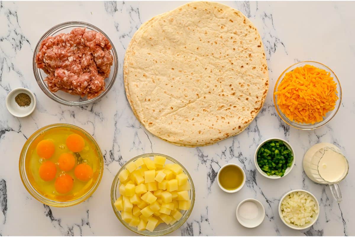 Tortillas, eggs, potatoes, cream, cheese, onions, peppers, and sausage on counter.