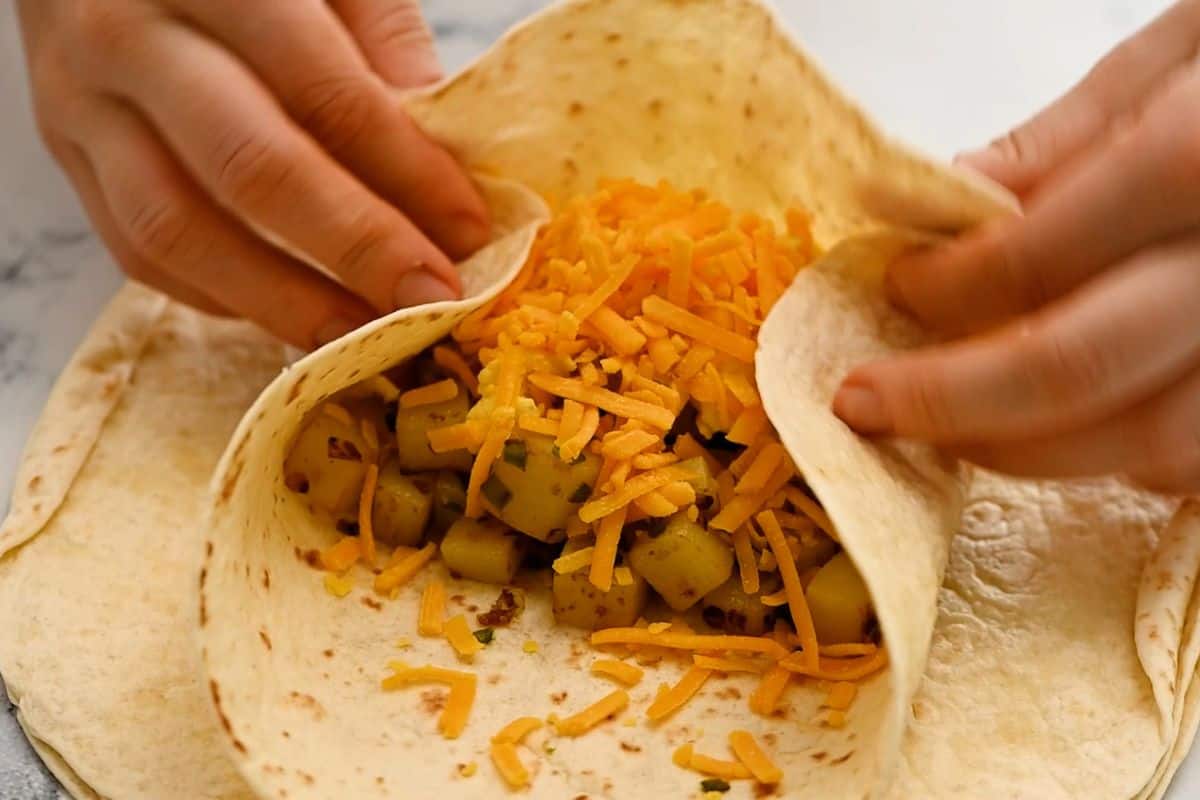 Flour tortilla filled with potatoes, eggs, sausage, and cheese being tucked and rolled.