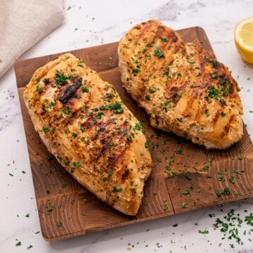 Cooked marinated Greek chicken on wooden cutting board topped with fresh parsley.