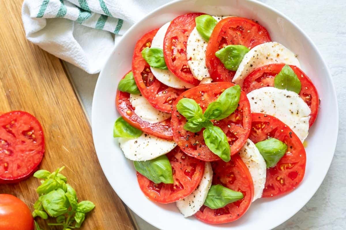 Alternating layers of tomatoes, mozzarella, and basil in shallow white serving platter topped with olive oil. next to cutting board with basil and tomatoes.