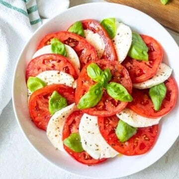 Alternating layers of tomatoes, mozzarella, and basil in shallow white serving platter topped with olive oil.