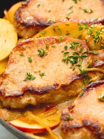 Close up of pork chops with apples in saute pan topped with fresh thyme leaves.