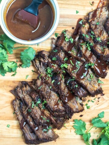 Sliced Instant Pot Brisket on wooden cutting board drizzled with BBQ sauce and parsley on the side.