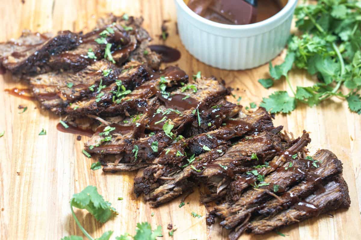 Sliced Instant Pot Brisket on wooden cutting board drizzled with BBQ sauce and parsley on the side.