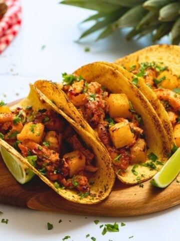 Chicken al pastor tacos on wooden tray with fresh lime wedges and minced cilantro.