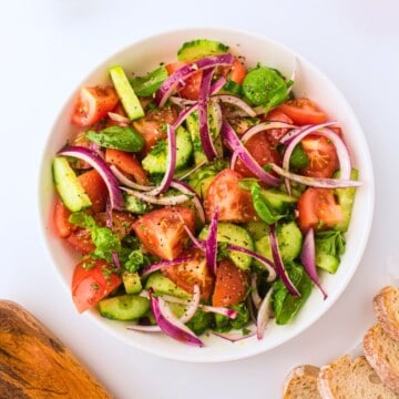 Tomato and cucumber salad with red onions, basil, and a Greek dressing in white bowl with fresh tomatoes in background.