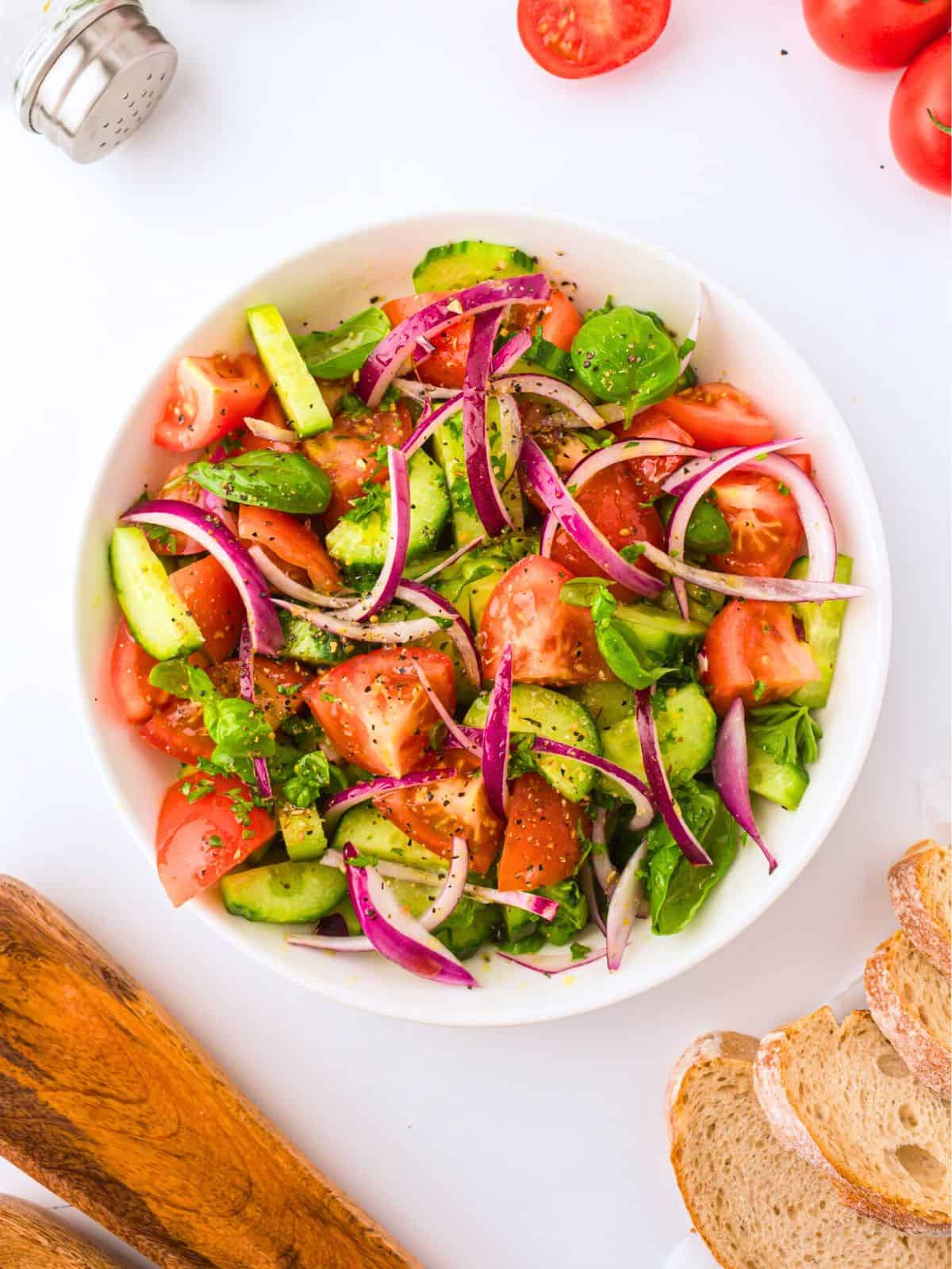 Sliced cucumbers, diced tomatoes, sliced red onions, and fresh basil tossed together with a simple Greek dressing in a white bowl.