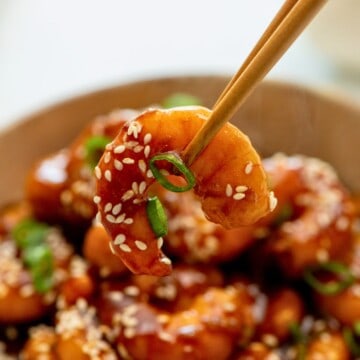 Chopstick holding one shrimp with bowl of General Tso Shrimp in the background.