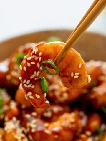 Chopstick holding one shrimp with bowl of General Tso Shrimp in the background.