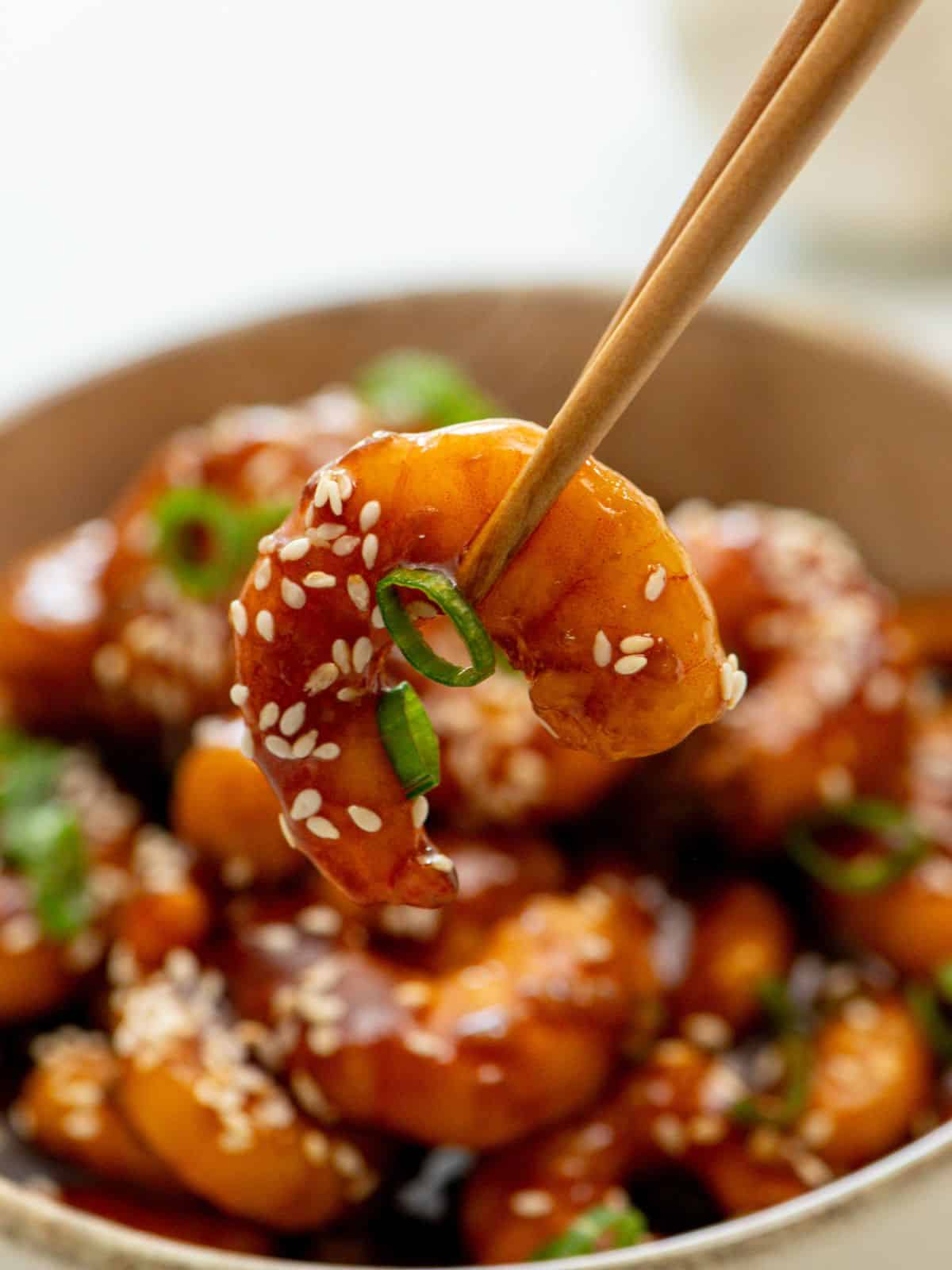 Chopstick holding one shrimp with bowl of General Tso Shrimp in the background.