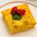 Slice of green chile egg casserole on white plate topped with diced tomatoes and cilantro.