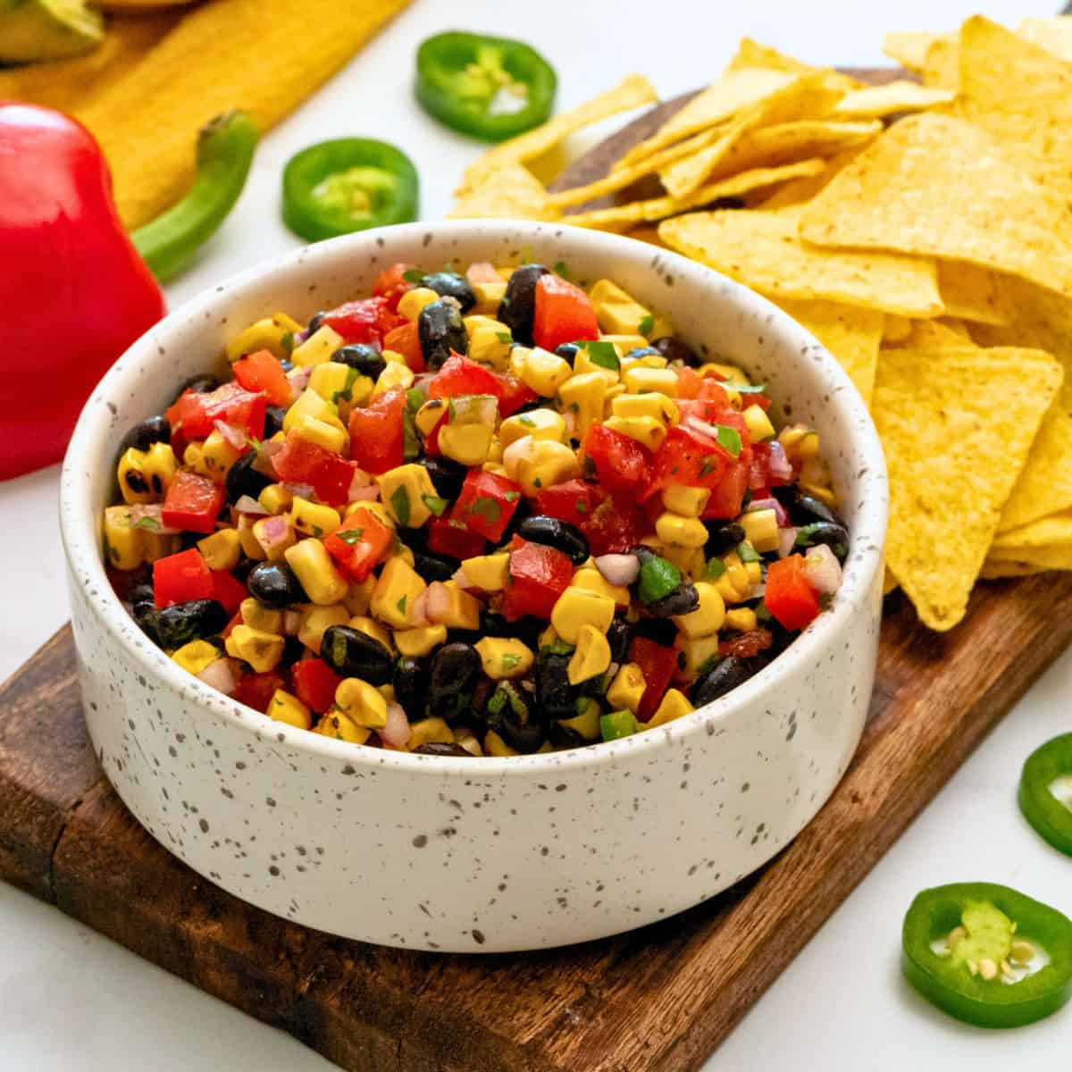 Easy Super Bowl Recipes Black bean and grilled corn tossed with tomatoes, peppers, jalapenos, and lime dressing in white bowl next to tortilla chips.