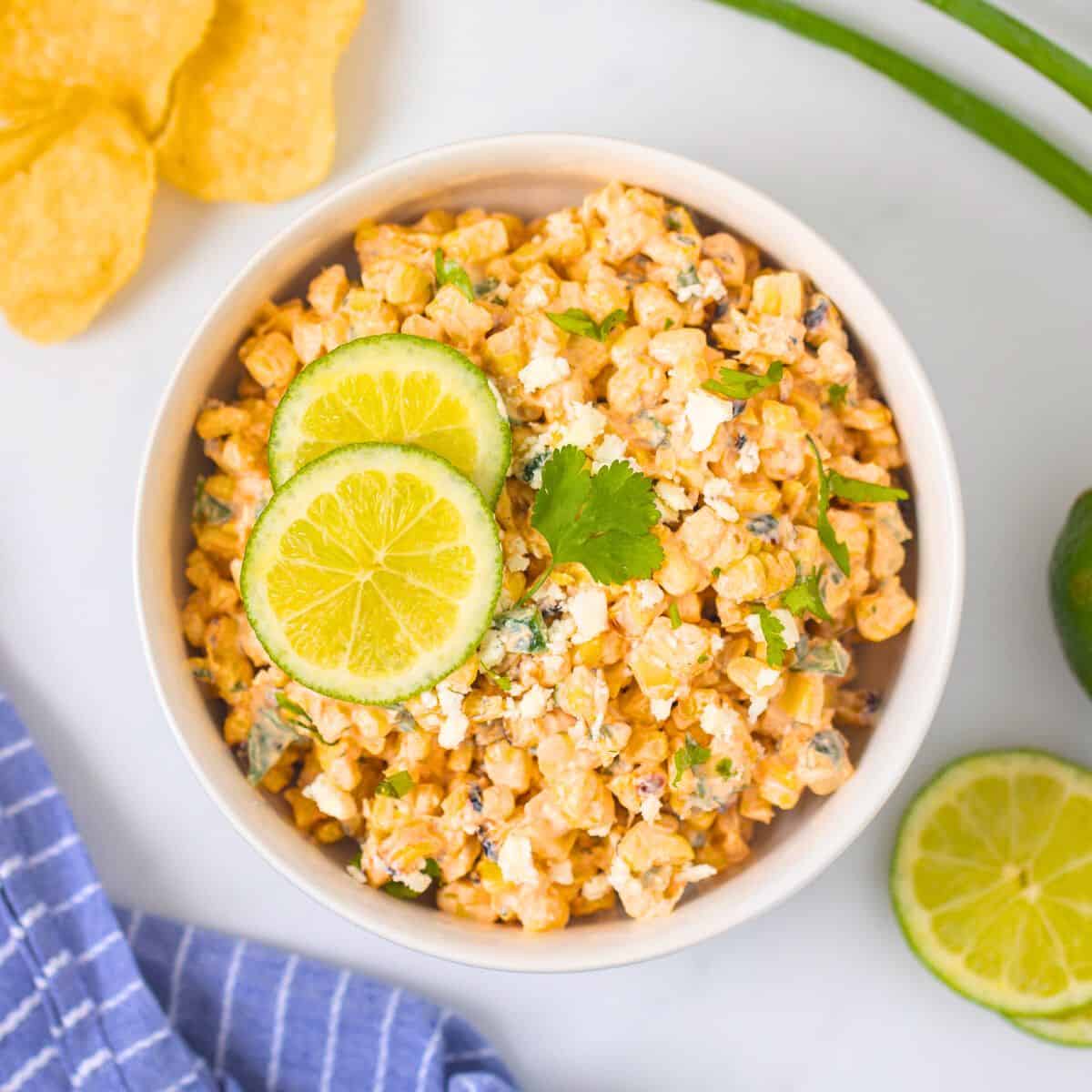 Super Bowl Party Recipes Mexican Street Corn Salad in white bowl topped with sliced limes, queso fresco, and cilantro.