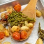 Wooden spoon scooping up roasted sheet pan gnocchi, tomatoes, and broccolini seasoned with Italian seasoning.