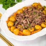 Shredded crockpot roast and potatoes and carrots on white platter next to parsley, horseradish, and gold serving spoons.
