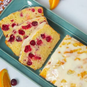 Cranberry orange bread with glaze on blue plate with a few slices cut to reveal fresh cranberries studding moist quick bread.