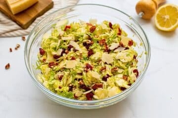Large mixing bowl with shaved Brussels Sprouts tossed with honey lemon dressing, diced apples, dried cranberries, shaved parmesan, and chopped toasted pecans.