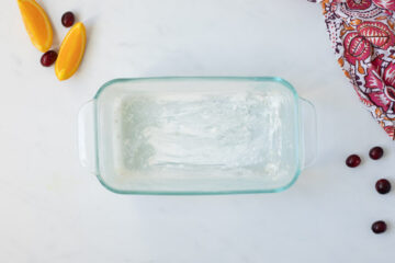 Buttered and flour dusted 8x4-inch glass loaf pan on counter next to fresh cranberries and orange slices.