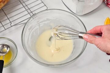 Wire whisk showing orange glaze dripping into mixing bowl with sugar and orange juice whisked together.