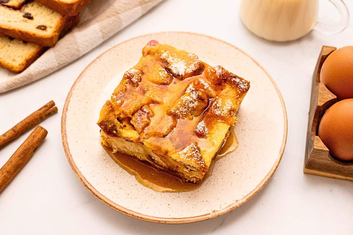 Make Ahead Easter Brunch Slice of overnight breakfast casserole made with cinnamon raisin bread, eggs, cubed ham, sharp cheddar, and eggs on plate topped with maple syrup and powdered sugar.