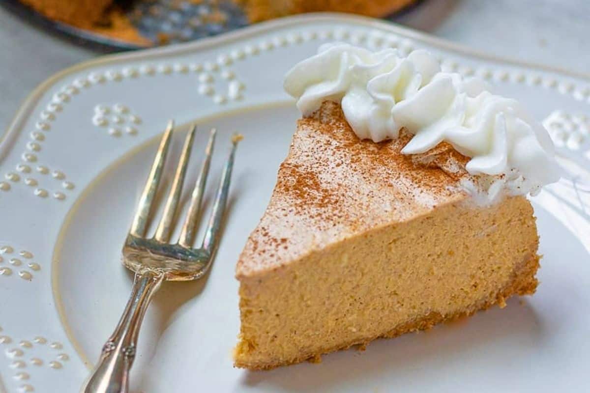 Make Ahead thanksgiving Recipes Instant Pot Slice of Instant Pot Pumpkin Cheesecake on plate with fork with remaining pumpkin cheesecake in background.