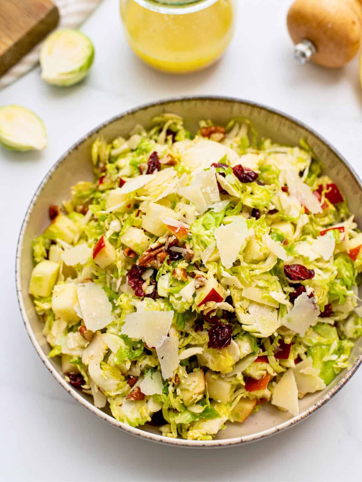 Shaved Brussels Sprouts Salad Shaved Brussels Sprouts tossed with honey lemon dressing, diced apples, dried cranberries, shaved parmesan, and chopped toasted pecans dished out in serving bowl with dressing in background.