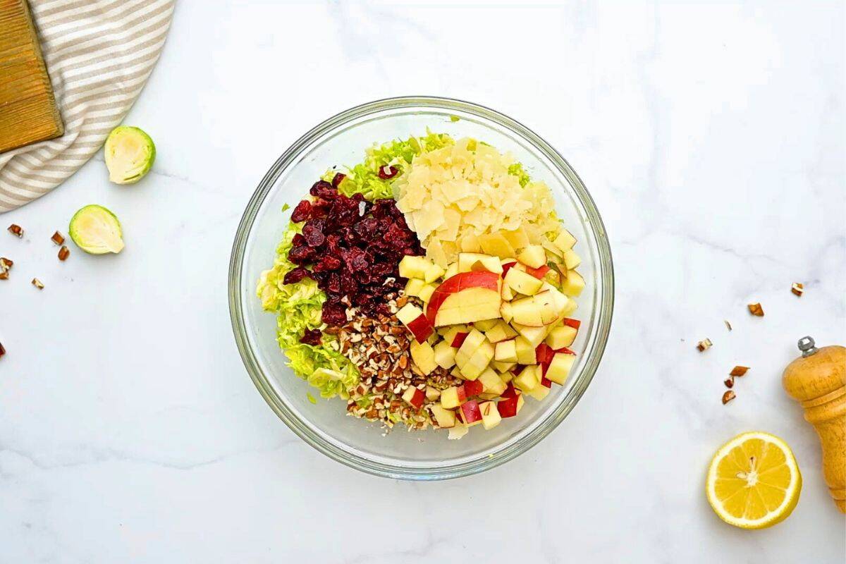 Easy Shaved Brussels Sprouts Salad Large mixing bowl with shaved Brussels Sprouts topped with diced apples, dried cranberries, shaved parmesan, and chopped toasted pecans.