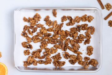 Walnuts coated with caramelized mixture made from orange juice, orange zest, brown sugar, white sugar, cinnamon, salt, and cayenne spread out on parchment paper lined baking sheet.