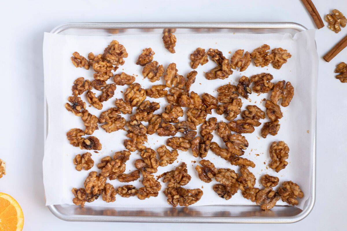 Walnuts coated with caramelized mixture made from orange juice, orange zest, brown sugar, white sugar, cinnamon, salt, and cayenne spread out on parchment paper lined baking sheet.