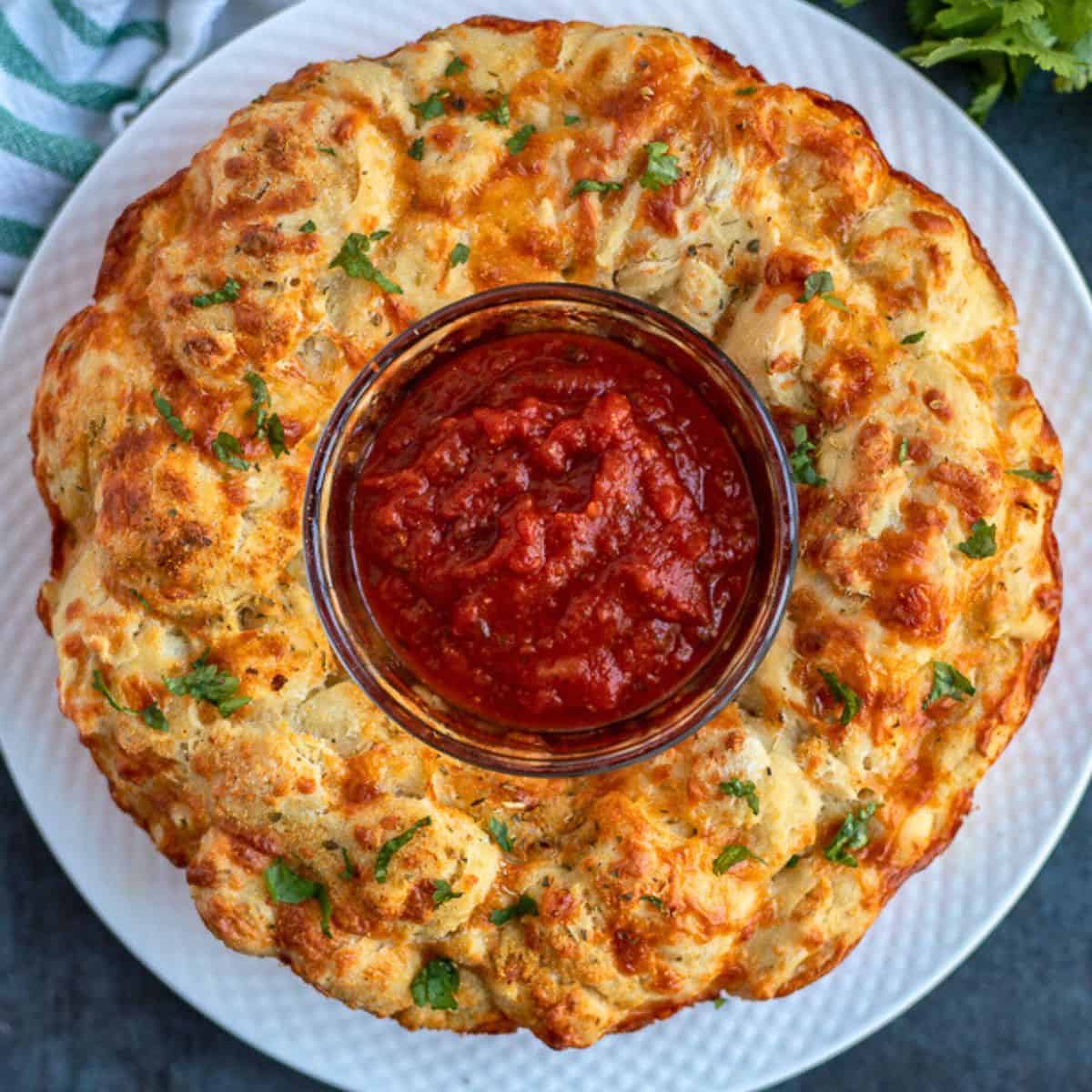 Game Day Recipes Pizza bread inverted on white plate topped with parsley and served with bowl of pizza sauce in middle of bread.