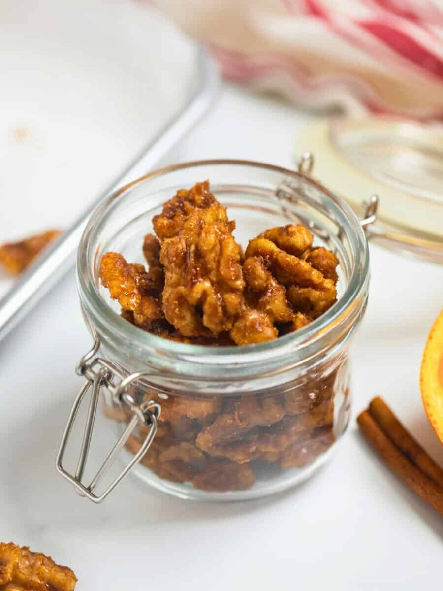 Small jar of homemade candied walnuts made with cinnamon, cayenne, and orange next to fresh orange and cinnamon sticks.