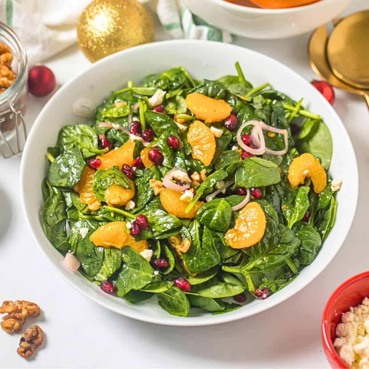 Christmas Dinner Recipes Salad made with baby spinach, mandarin oranges, pomegranate arils, candied walnuts, crumbled feta, shallots, and champagne vinaigrette in white bowl next to candied nuts and shallots.