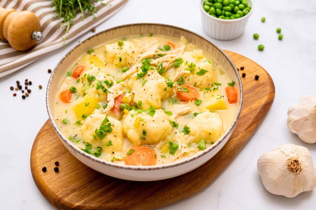 Bowl of creamy soup with shredded chicken, cooked carrots, potatoes, peas and homemade dumplings topped with cracked black pepper and fresh parsley.