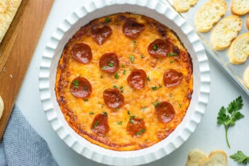 Baked pizza dip in white pie plate topped with pepperoni and parsley and served with toasted crostini.