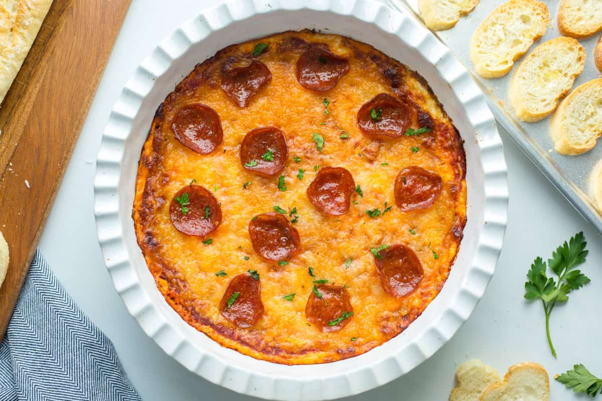 Baked pizza dip in white pie plate topped with pepperoni and parsley and served with toasted crostini.