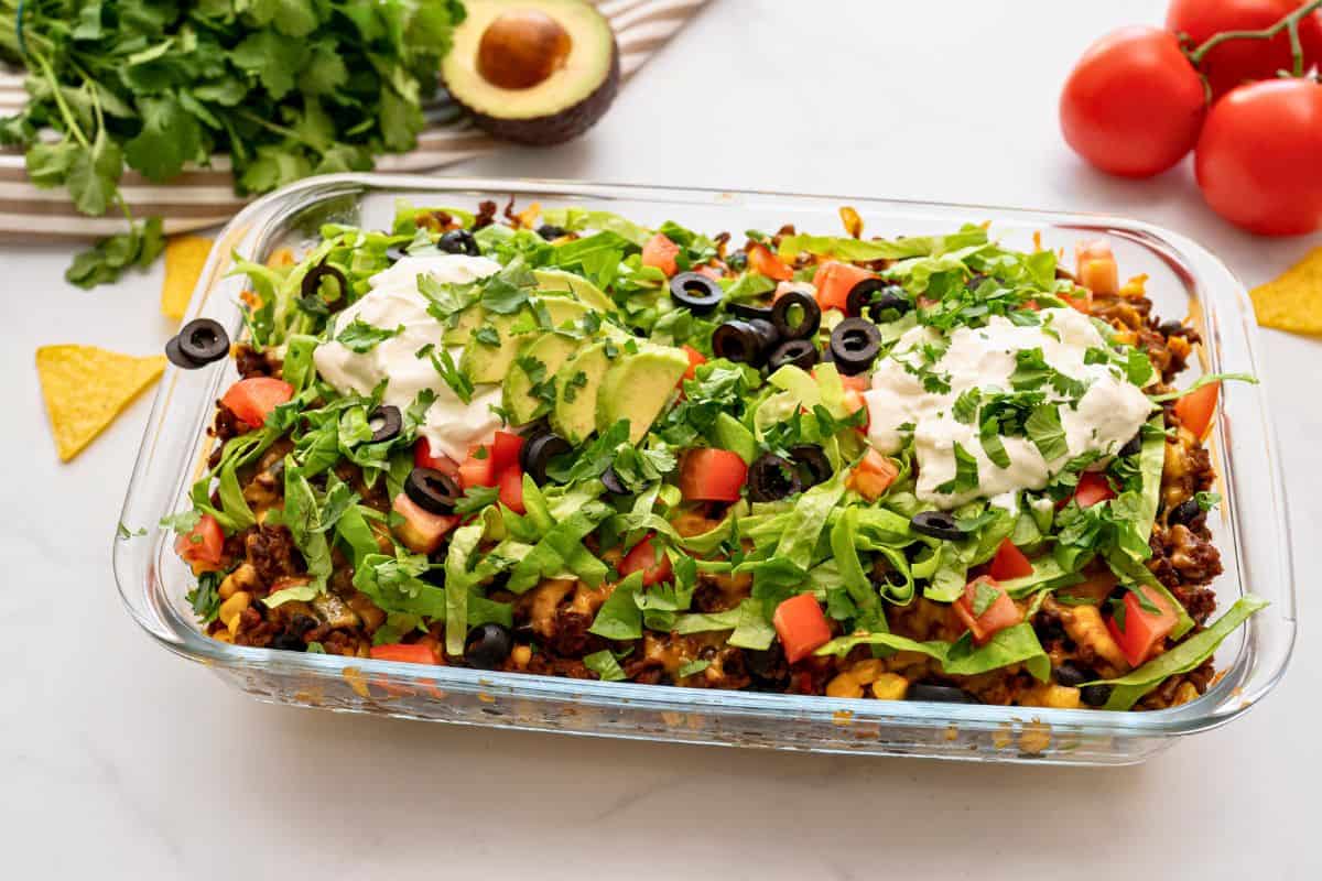 Taco Bake Recipe Tortilla chips, beef, bean, and corn mixture layered in casserole dish topped with cheese and baked then topped with lettuce, tomatoes, olives, sour cream and avocado.