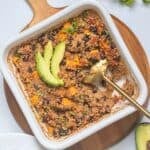 Baked quinoa with sweet potatoes, black beans, and diced tomatoes in white dish topped with avocado and cilantro with gold serving spoon taking out some.