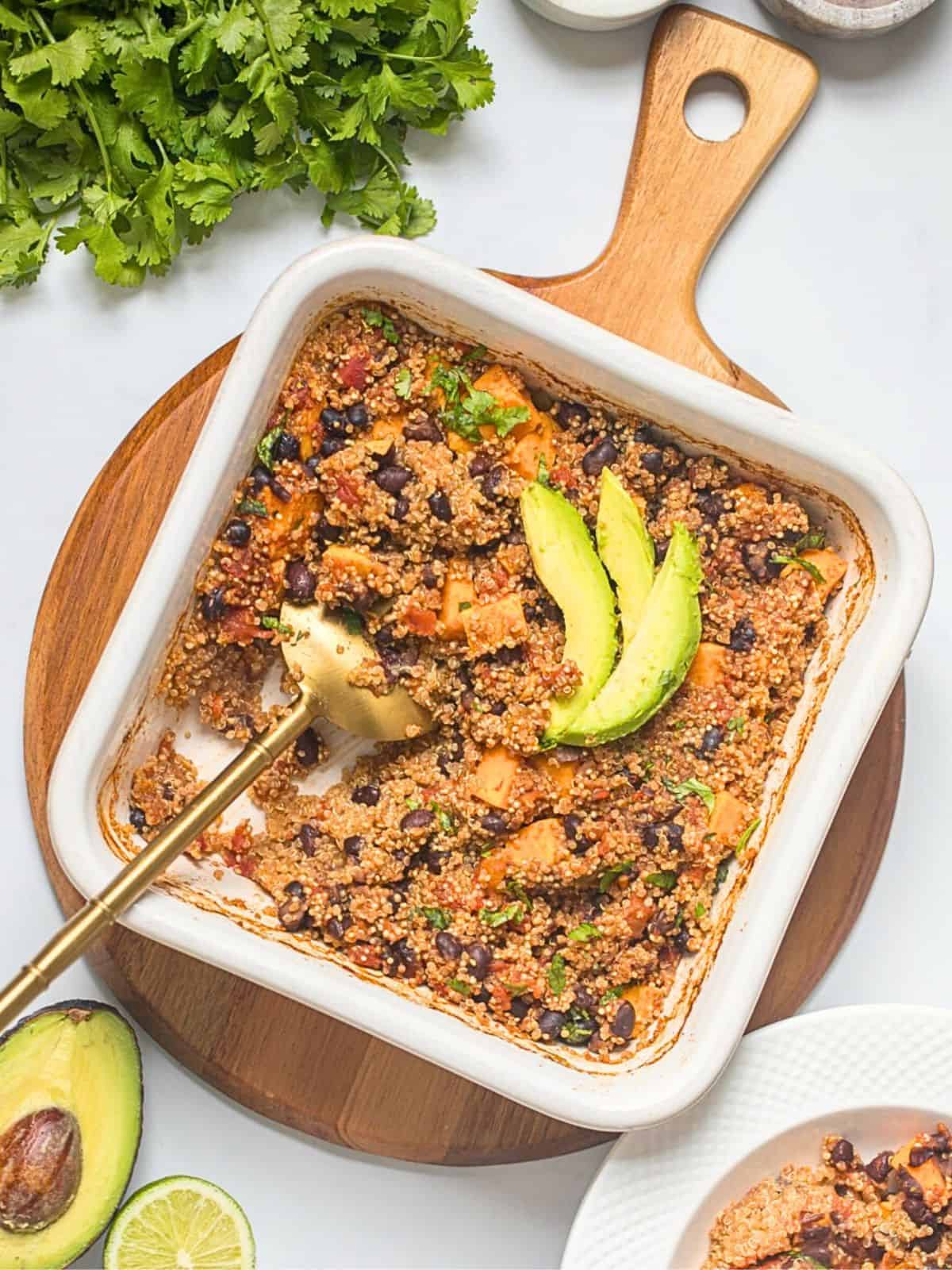 Baked quinoa with sweet potatoes, black beans, and diced tomatoes in white dish topped with avocado and minced cilantro and lime on the side.